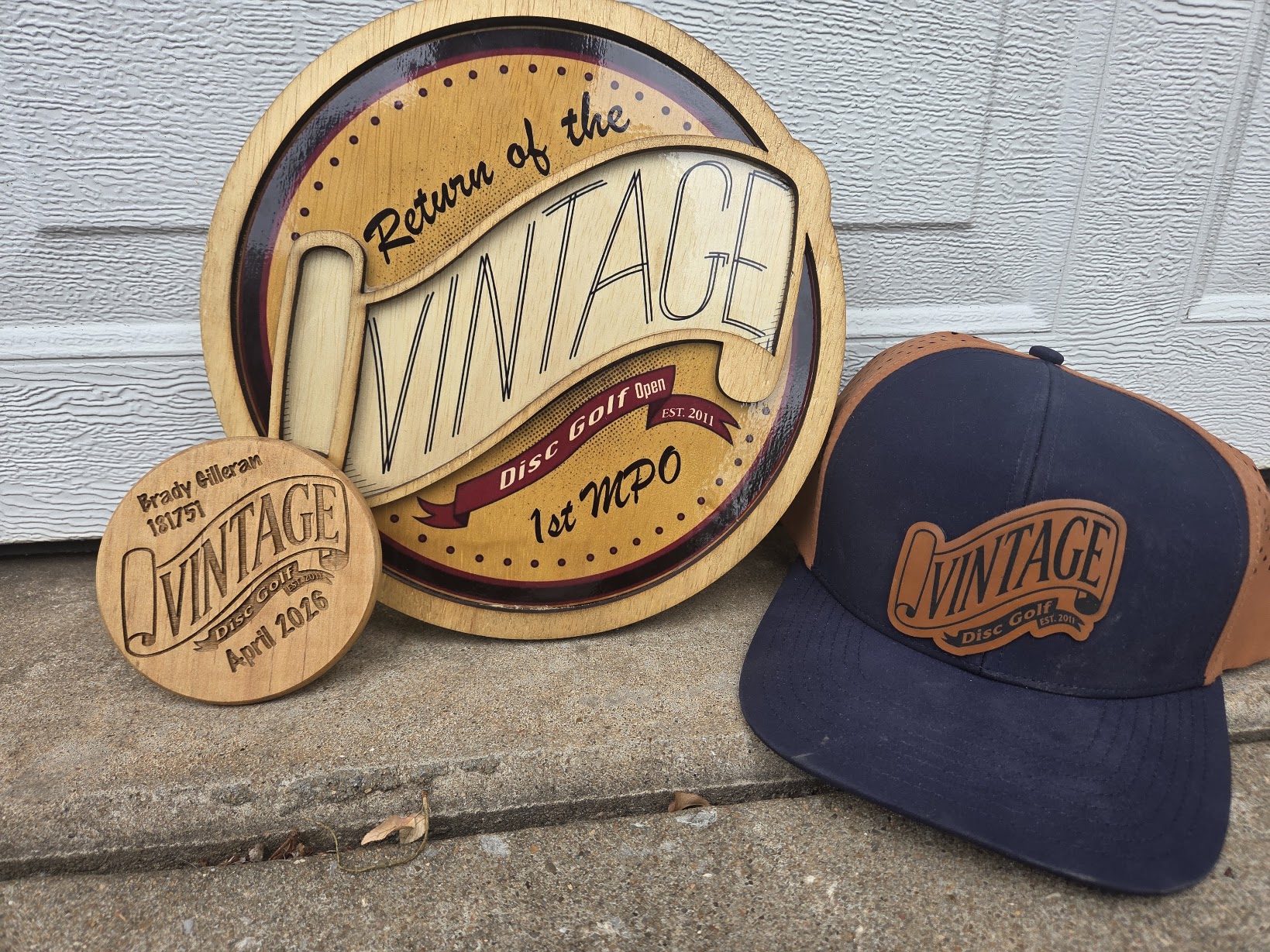 A layered wooden Vintage Disc Golf Open plaque, an engraved wooden mini, and a navy hat with a leather patch